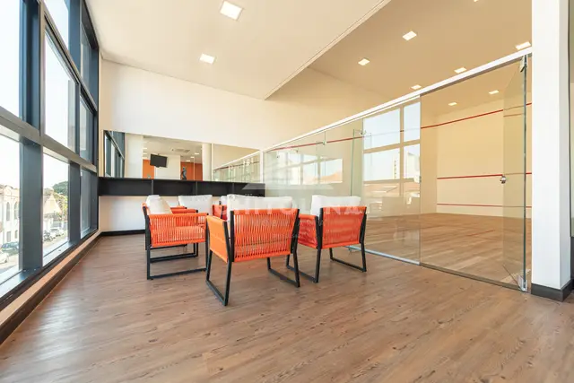 Quadra de squash com parede de vidro no Edifício Cathedral e estar com poltronas laranja ao lado.